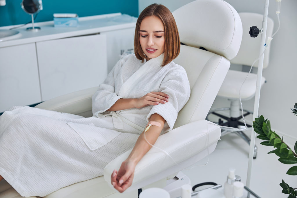 Waist up side view portrait of redheaded Caucasian female in white bathrobe sitting in medicine armchair during medical procedure in wellness center