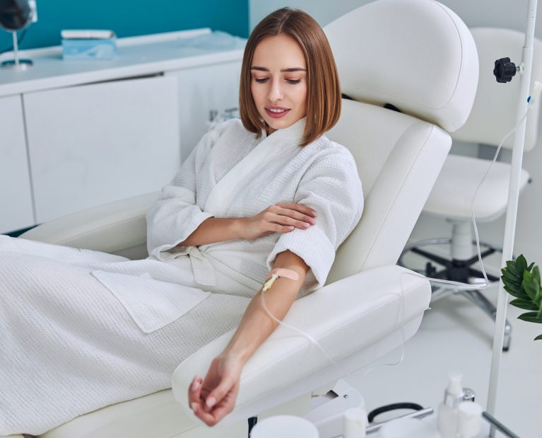 Waist up side view portrait of redheaded Caucasian female in white bathrobe sitting in medicine armchair during medical procedure in wellness center
